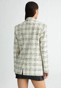 Blazer à carreaux en tissu crème avec un motif de grille noire. Il possède une fente unique à l'arrière et une coupe ajustée. Longueur courte.