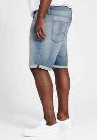 Pantalones cortos de mezclilla azul claro con puños enrollados, que cuentan con un diseño de cinco bolsillos y un ligero desvanecimiento. Combinados con zapatillas blancas. Vista trasera.