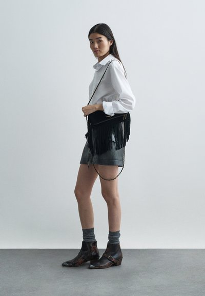 Melnas ādas plecu soma ar fringing detalēm, kopā ar baltu krekls, pelēkiem šortiem un teksturētām potīšu zābakiem. Neitrāla fona.