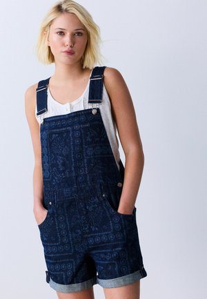 Junge Frau mit blondem Haar, die ein weißes ärmelloses Top und dunkelblaue gemusterte kurze Denim-Latzhosen trägt, Hände in den Taschen.