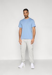 Μπλε βαμβακερό t-shirt με στρογγυλή λαιμόκοψη και λογότυπο, συνδυασμένο με γκρι φόρμες που διαθέτουν τσέπες cargo και μανσέτες, ολοκληρωμένο με λευκά αθλητικά παπούτσια.