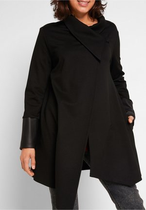 Kobieta w długim czarnym płaszczu z detalami ze skóry na mankietach, stojąca z rękami w kieszeniach, uśmiechająca się na białym tle.