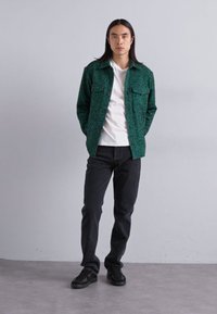 Green patterned jacket with two chest pockets, worn over a white T-shirt and paired with black jeans and sneakers. Model stands relaxed.