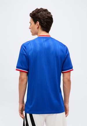 Giovane uomo con capelli scuri corti che indossa una camicia blu a maniche corte con rifiniture rosse e bianche, volto di schiena su uno sfondo semplice.