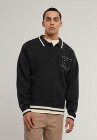 Černý pletený svetr s bílým proužkovaným límcem a manžetami, na levé části hrudi s písmenem "K" a vzorem hvězd.