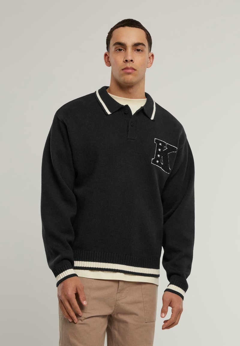 Černý pletený svetr s bílým proužkovaným límcem a manžetami, na levé části hrudi s písmenem "K" a vzorem hvězd.