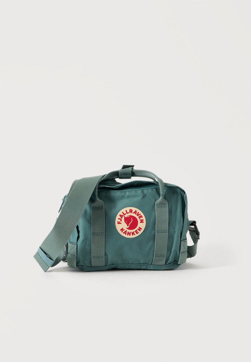 Pequeña mochila verde Fjällräven Kånken con correa de hombro ajustable y parche con el logo en la parte delantera sobre un fondo liso.