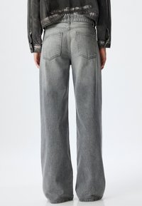 Jeans de pierna ancha grises, hechos de tela de mezclilla, con una textura desgastada. Presentan dos bolsillos traseros y una cinturilla estándar.