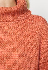 Gros plan sur une personne portant un pull à col roulé en tricot épais et duveteux de couleur orange, sur un fond clair uni.