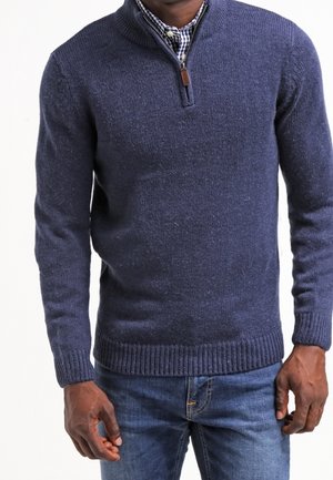 Homme portant un pull marine à fermeture éclair jusqu'au quart sur une chemise à carreaux noir et blanc avec un jean bleu, debout les bras détendus.