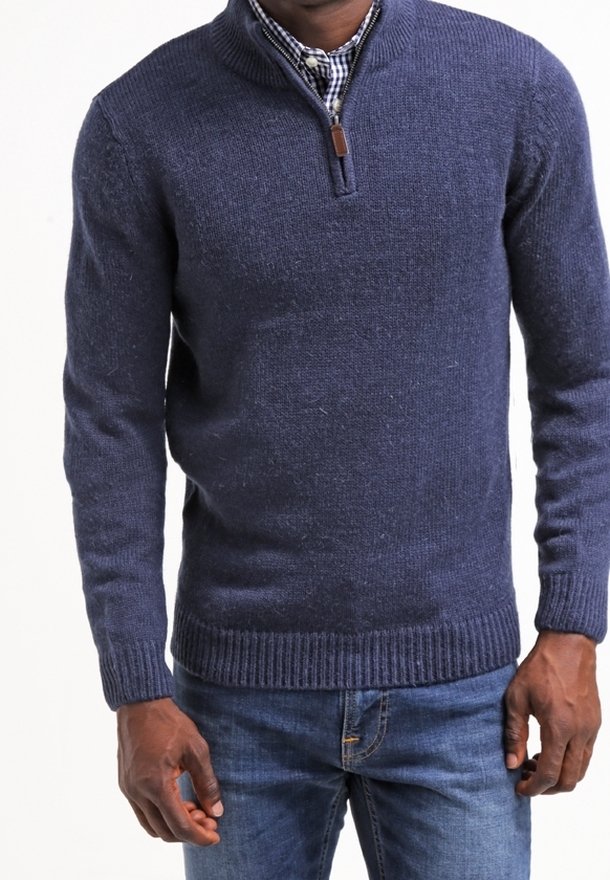 Homme portant un pull marine à fermeture éclair jusqu'au quart sur une chemise à carreaux noir et blanc avec un jean bleu, debout les bras détendus.