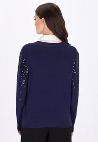 Dunkelblauer Pullover mit geripptem Struktur und mit Pailletten verzierten Ärmeln. Rundhalsausschnitt mit einem kontrastierenden weißen Kragen darunter.