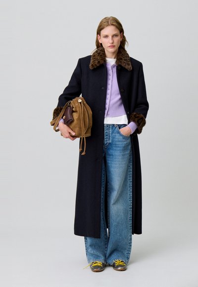 Largo abrigo negro con cuello y puños de estampado de leopardo, superpuesto a un cárdigan lavanda y una camisa blanca, combinado con jeans azules sueltos y un bolso marrón.