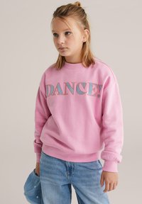 Plavolaska deklica v roza majici z napisom "DANCE!", modrih kavbojkah, drži modro kapico in gleda vstran na enobarvnem ozadju.