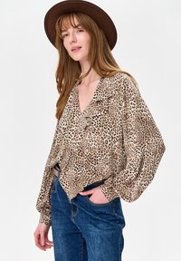 Kobieta w brązowym kapeluszu, bluzce w panterkę z długim rękawem i falbankami oraz niebieskich dżinsach, pozująca z jedną ręką w kieszeni na białym tle.