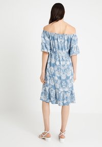 Nainen, jolla on päällään sininen off-shoulder kukkakuosiset mekko, jossa on röyhelöhiha ja helma, yhdistettynä valkoisiin remmillä varustettuihin paksupohjaisiin sandaaleihin, kääntyi pois.