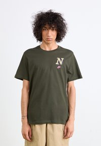 Oliven grøn t-shirt i bomuld med korte ærmer, med et mønstret "N" logo og pink Nike swoosh på brystet. Lige snit i kanten.