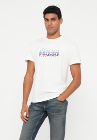 Άνδρας που φοράει λευκό κοντομάνικο μπλουζάκι με το λογότυπο "1985 Tommy Jeans" και μπλε τζιν, που στέκεται μπροστά σε απλό λευκό φόντο.