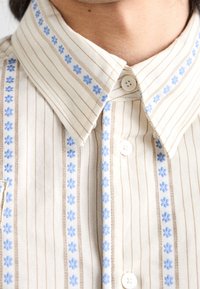 Collared shirt with thin beige stripes, featuring blue floral embroidery along the button placket and collar. Light fabric texture.