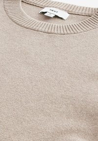 Beżowy sweter dzianinowy z okrągłym prążkowanym dekoltem. Materiał ma teksturowany splot, co nadaje mu miękki i ciepły wygląd. Wewnątrz widoczna metka.