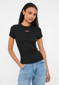 Musthave lühikeste varrukatega liibuv ribidega must t-särk, millel on ümar kaelus ja roosa brändi logo rindkerel. Kangas näeb välja sile ja veniv.