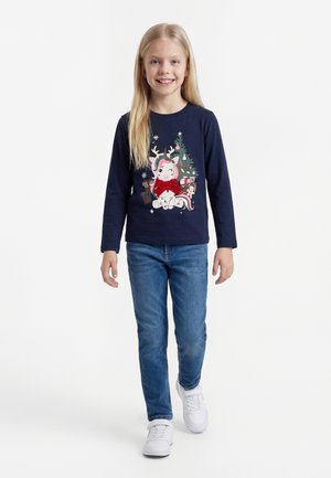 Smilende pige går, iført hvide sneakers, blå jeans og en marinblå langærmet skjorte med et festligt enhjørninge- og juletræsdesign.