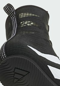 Close-up af en sort sportsko med tekstureret sål, hvide striber på siden og en pull-tab med hvid søm ved hælen.