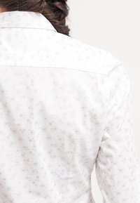Femme portant une chemise blanche avec de petits motifs floraux gris clair, cheveux tressés visibles de dos.