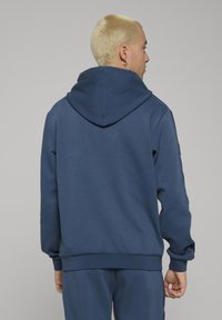 Navy zila kapuce ar rāvējslēdzēju ar vaļēju piegriezumu, ribota aproce un apkakle. Gluda tekstūra un izteiksmīga kapuce ar centrālo šuvi.