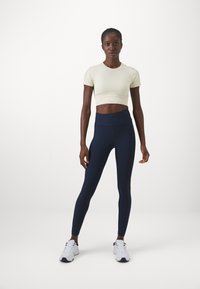 Svijetlosmeđi kratki crop top s kratkim rukavima i rebrastom teksturom, u kombinaciji s visokim mornarskim tajicama. Oboje pružaju pripijeni sportski izgled.