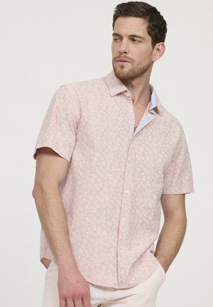 Un homme portant une chemise à manches courtes rose clair à boutons avec de petits motifs floraux blancs, regardant sur le côté avec une main dans sa poche.