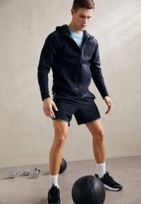 Completo sportivo con una felpa con zip blu navy, maglietta azzurro chiaro, pantaloni corti neri e sneakers nere, ambientato su uno sfondo neutro.