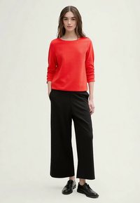 Raudonas faktūrinis marškinėliai ilgomis rankovėmis, derinami su juodomis plačiomis kelnėmis ir juodais bateliais su raišteliais, pateikti prieš neutralią foną.