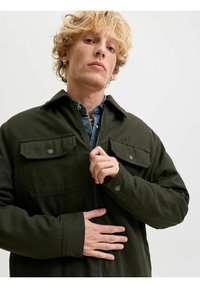Jeune homme blond portant des boucles d'oreilles créoles dorées ferme une veste vert foncé par-dessus une chemise en jean sur un fond clair uni.