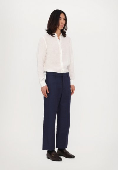 Fehér vászon ing, sötétkék nadrág és sötétbarna mokaszin. Kicsit texturált anyaggal és laza fit-tel rendelkezik, egyenes lábszárral.