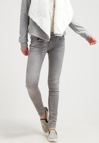 Jeans skinny grises con un acabado desgastado, combinados con una chaqueta de forro polar gris claro, que presenta un cuello ancho. Unos zapatos ligeros y de fácil puesta completan el conjunto.