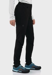 Schwarze Sporthosen mit einer schmalen Passform, ausgestattet mit einer Seitentasche und elastischen Bündchen. Kombiniert mit blauen und schwarzen Multifunktionsschuhen.