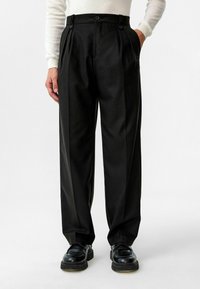 Zwarte geplooide broek met een getailleerde snit, gemaakt van een gladde stof, voorzien van twee voorkantzakken en een hoge taille met knoopaccenten.
