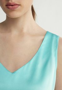 En närbild av en ljus turkos satinplagg med V-ringad halslinje och slät yta, som visar en subtil glans och vinklade axelremmar.