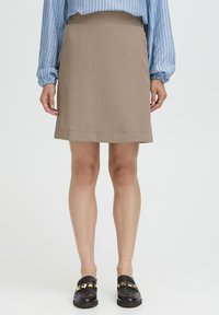 Mujer con una falda beige hasta la rodilla, blusa de rayas azul de manga larga y zapatos de charol negros con adornos cuadrados dorados, de pie sobre un suelo blanco.