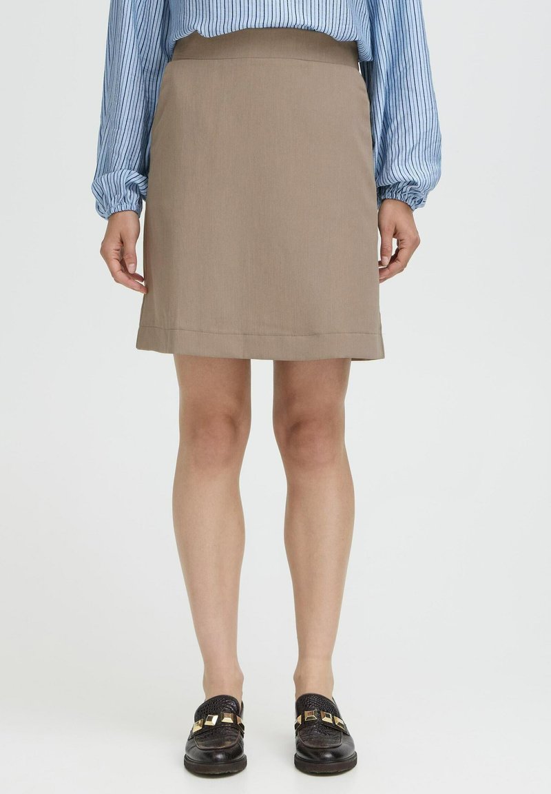 Mujer con una falda beige hasta la rodilla, blusa de rayas azul de manga larga y zapatos de charol negros con adornos cuadrados dorados, de pie sobre un suelo blanco.