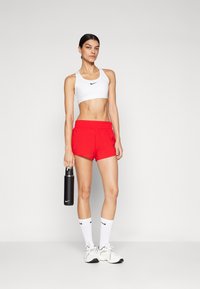 Reggiseno sportivo Nike bianco, pantaloni corti rossi, bottiglia d'acqua nera, calze bianche e sneaker grigi. Presenta un cinturino elastico e dettagli con logo.