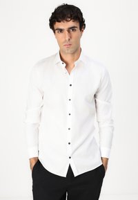 Hvit langermet skjorte laget av et lett stoff med svarte knapper foran og klassisk krage. Ren, skreddersydd design.