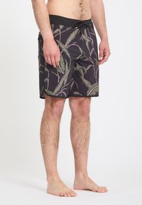 Homme portant un short de bain noir avec un motif de feuilles vertes et roses, debout pieds nus devant un fond uni.