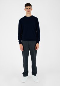 Granatowy dziany sweter, szare spodnie o subtelnej fakturze i białe sneakersy. Model stoi na tle jednolitego białego tła.