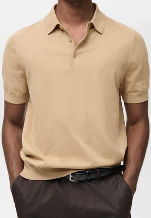 Polo beige a maniche corte indossata con pantaloni scuri e cintura nera, mani nelle tasche, primo piano che mostra la parte superiore del torso e i dettagli del colletto.
