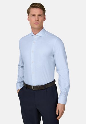 Mężczyzna ubrany w jasnoniebieską koszulę wpuszczoną w ciemnogranatowe spodnie, z czarnym paskiem, stojący na jasnym, jednolitym tle.