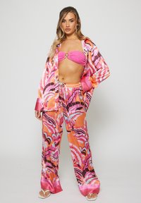 Una joven se encuentra vestida con un top bandeau de color rosa brillante, pantalones de seda naranja y rosa con un gran estampado de flores y una camisa abierta a juego.