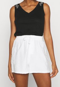 Svart ärmlös topp med V-ringning och prydnadsdetaljer på axlarna, kombinerad med vita shorts med elastisk midja och sidofickor.