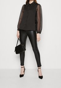 Svart blus med genomskinliga ärmar och nitar på kragen, kombinerad med glänsande svarta leggings och svarta klackar. Modellen håller en liten svart handväska.